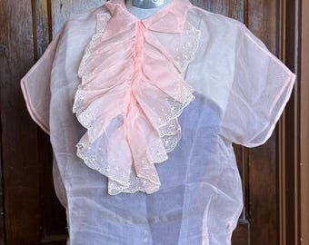 May include: A sheer, light pink blouse with short sleeves and a ruffled front. The ruffle is a darker shade of pink and has delicate white embroidered details. The blouse is displayed on a mannequin, showcasing its vintage style.