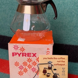 May include: A vintage Pyrex coffee pot with a gold-colored band and black handle, sitting on top of its original orange box. The box features floral designs and the Pyrex logo. A small sign reads "you taste the coffee... not the coffeepot..."