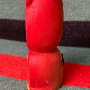 May include: A bright red ceramic vase shaped like a stylized character, featuring a smooth matte finish, a hollow top opening, and a small dark circular detail on the side. The object has a rounded upper section and a slightly wider base.