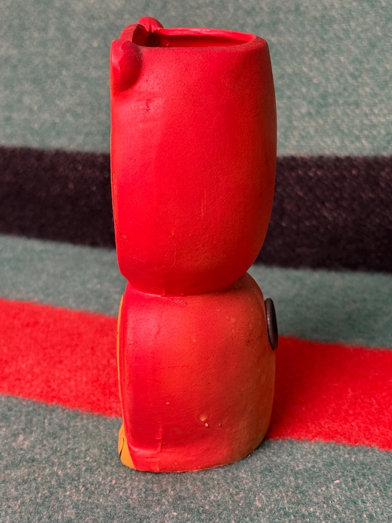 May include: A bright red ceramic vase shaped like a stylized character, featuring a smooth matte finish, a hollow top opening, and a small dark circular detail on the side. The object has a rounded upper section and a slightly wider base.