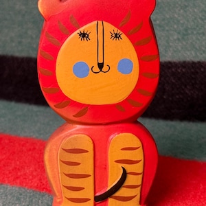 May include: A hand-painted wooden figurine of a lion with a bright red mane, a yellow face, and brown stripes. The lion has blue circular cheeks, black painted eyes with lashes, and a black nose and mouth. The body is red with yellow legs.