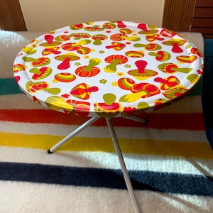 May include: A white metal table with a round top featuring a psychedelic mushroom pattern in shades of red, yellow, and green. The table has three legs.