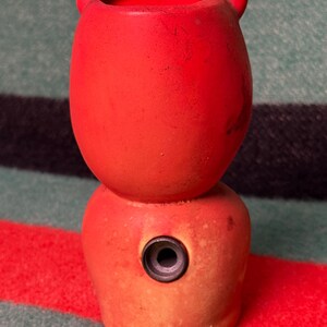 May include: A bright red, two-tiered ceramic vessel with a rounded top section featuring two small ear-like protrusions and a wider base section. The lower portion includes a single, centered circular opening with a dark grey rim.