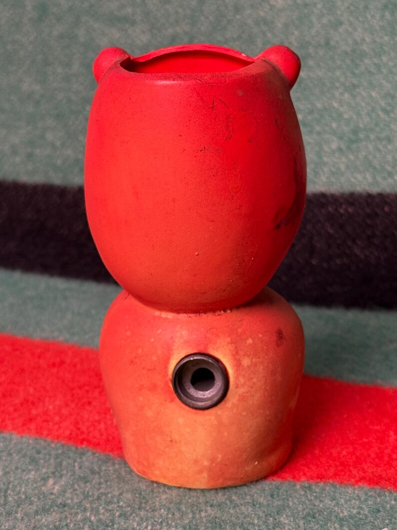 May include: A bright red, two-tiered ceramic vessel with a rounded top section featuring two small ear-like protrusions and a wider base section. The lower portion includes a single, centered circular opening with a dark grey rim.