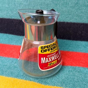 May include: A vintage glass coffee carafe with a metal lid and handle. The carafe has a clear body with a label that reads "Instant Maxwell House Coffee 10 oz." and a "Special Offer" sticker. The carafe is empty.