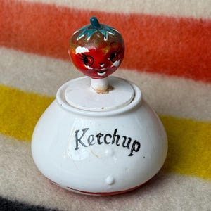 May include: A white ceramic ketchup dispenser with a red and green tomato-shaped lid. The lid has a painted face with black eyes and a smile. The dispenser says "Ketchup" in black lettering.