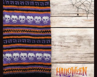 May include: A black, orange, and purple knitted scarf with a pattern of white skulls. The scarf is on a wooden background with a spider web and the word "HALLOWEEN" in orange and purple letters.