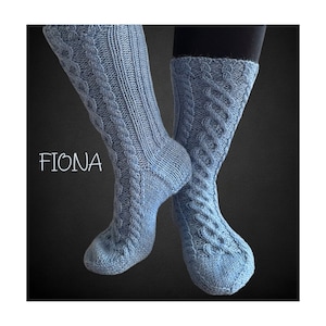 Peut inclure: Une paire de chaussettes bleues en tricot avec un motif en câble. Les chaussettes sont présentées sur les jambes d'une personne. Le mot "FIONA" est écrit en blanc sur le côté gauche de l'image.