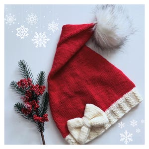 May include: Red knitted Santa hat with a white pom pom and a white bow. The hat is on a white background with a sprig of greenery.