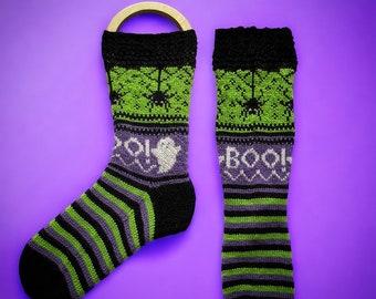 May include: A pair of black, green, and purple knitted socks with a Halloween theme. One sock has a ghost and the word "Boo!" on it, while the other sock has a spider web and a spider on it.