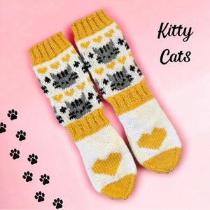 Puede incluir: Un par de calcetines de punto con un diseño de gato y corazón. Los calcetines son blancos con detalles amarillos y grises. El texto "Kitty Cats" se muestra sobre un fondo rosa.