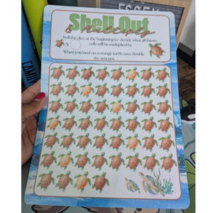 May include: A rectangular game board titled Shell Out & Multiply with a grid of small brown and orange turtle illustrations. The text reads: Roll the dice at the beginning to decide what all future rolls will be multiplied by. X [circle] When you land on an orange turtle save double the amount.