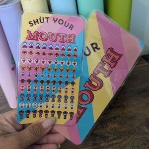 May include: A set of two colourful stickers with the phrase "Shut Your Mouth" printed on them. The stickers feature a variety of cartoon mouths with different expressions.
