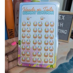 May include: A clear plastic card with the text "Heads or Tails BUNNY EDITION" in blue and green. The card is filled with small bunny and basket stickers in various colours. The card is held by a person with pink nail polish.
