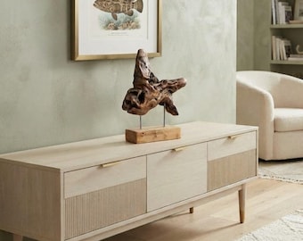 May include: A light-toned wooden media console featuring three cabinet doors with vertical ribbed detailing, slim gold-toned handles, and tapered legs with gold-toned caps. A natural driftwood sculpture sits on top, and a framed fish print hangs above.