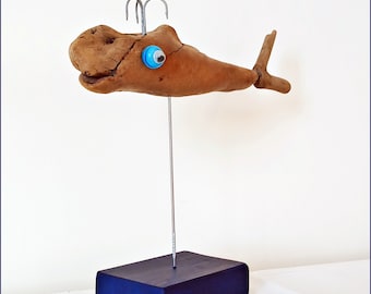 May include: A wooden whale sculpture with a blue eye, mounted on a dark blue wooden base. The whale is made from driftwood and has a whimsical, handcrafted look.