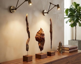 May include: Two brass wall-mounted swing-arm lamps with cylindrical heads illuminate three vertical wooden sculptures on square bases. The sculptures are arranged on a dark wood sideboard next to two stacked books and a stone obelisk.