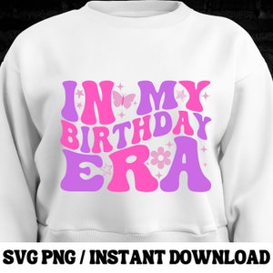 Puede incluir: Sudadera blanca con el texto "In My Birthday Era" en letras rosas y moradas con detalles de mariposas y flores.
