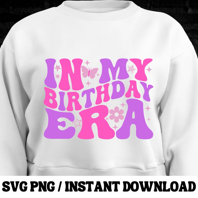 Puede incluir: Sudadera blanca con el texto "In My Birthday Era" en letras rosas y moradas con detalles de mariposas y flores.