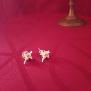 May include: A pair of unusual earrings, each with a small, off-white, star-shaped design and a central post. The earrings are set against a deep red background, with a brass candlestick visible in the background.