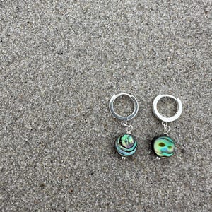 May include: Two silver hoop earrings, each with a round abalone shell bead. The beads display iridescent colours. The earrings are set against a textured, grey background. The earrings are a jewellery item.