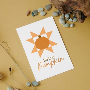 Puede incluir: Una tarjeta blanca con un diseño geométrico de calabaza en tonos naranja y marrón. El texto "Hello Pumpkin" está escrito en una fuente de escritura. La tarjeta está sobre una superficie beige con elementos decorativos.