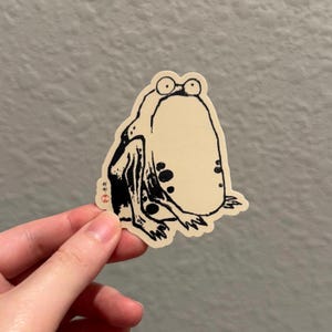 May include: Black and white line art sticker of a frog with large eyes and a round body. The sticker is on a beige background.