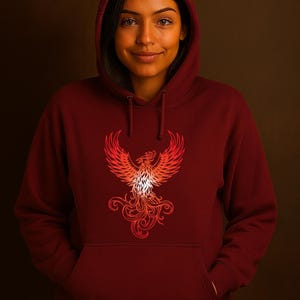 A woman stands against a deep brown background wearing a maroon hoodie with the hood up. The front of the hoodie showcases a bright red and orange phoenix graphic with detailed feathers and swirling flames. She has a gentle smile and her hands are tucked into the front pocket, exuding warmth and confidence. The lighting highlights her features and the bold contrast of the phoenix design against the dark hoodie fabric.