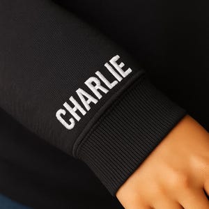 A close-up view of a black hoodie sleeve featuring the name "CHARLIE" embroidered in bold, white, all-caps block letters. The embroidery is placed just above the ribbed cuff. The image highlights the clean stitching and thick thread texture of the personalized design, as well as the smooth knit fabric of the hoodie. A hand rests naturally below the cuff, adding a sense of scale and wearability.