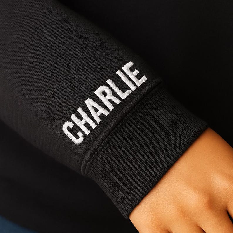A close-up view of a black hoodie sleeve featuring the name "CHARLIE" embroidered in bold, white, all-caps block letters. The embroidery is placed just above the ribbed cuff. The image highlights the clean stitching and thick thread texture of the personalized design, as well as the smooth knit fabric of the hoodie. A hand rests naturally below the cuff, adding a sense of scale and wearability.