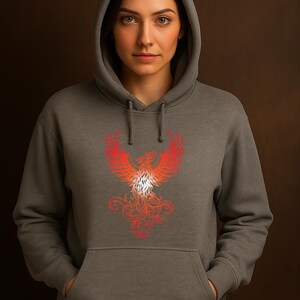 A woman wearing a dark gray hoodie stands confidently against a rich brown backdrop. The hoodie features a vibrant red and orange phoenix design on the front, with detailed feathering and swirling flames, symbolizing strength and rebirth. The hood is up, framing her face, and her hands are casually tucked into the front pocket. Her serious, direct gaze adds to the bold and empowering tone of the image.
