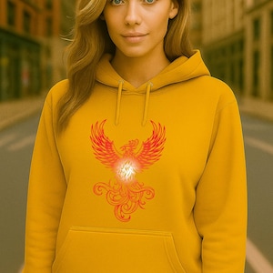 A young woman stands on a city street wearing a vibrant mustard yellow hoodie. The hoodie features a glowing red and orange phoenix graphic on the front, symbolizing fire and renewal. On the right sleeve cuff, the word "Phoenix" is embroidered in white block letters. The woman has long, wavy blonde hair and a soft smile, and the urban background is softly blurred to keep focus on her and the hoodie design.