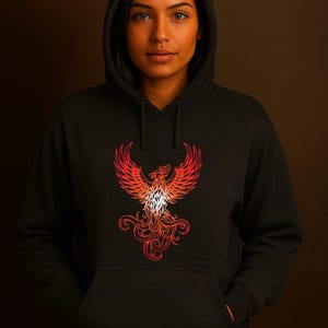 A woman stands against a dark brown background wearing a black hoodie with the hood up. The hoodie features a vivid red and orange phoenix graphic on the front, symbolizing rebirth and strength. Her hands are tucked into the front pocket, and she gazes directly at the camera with a calm, confident expression. The lighting highlights her warm skin tone and the intricate flame-like details of the phoenix design.
