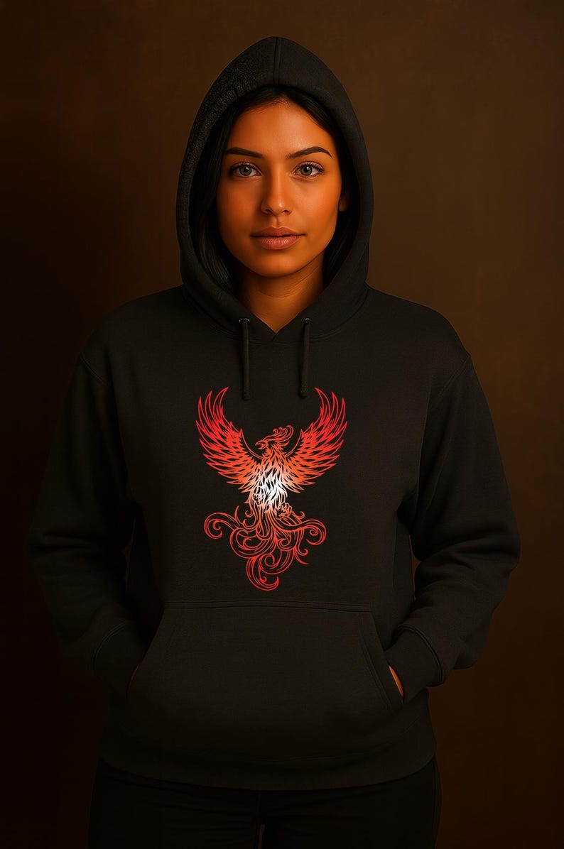 A woman stands against a dark brown background wearing a black hoodie with the hood up. The hoodie features a vivid red and orange phoenix graphic on the front, symbolizing rebirth and strength. Her hands are tucked into the front pocket, and she gazes directly at the camera with a calm, confident expression. The lighting highlights her warm skin tone and the intricate flame-like details of the phoenix design.