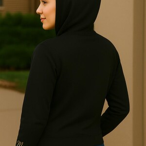A woman is shown from behind wearing a black hoodie with the hood up. The focus is on the right sleeve, which features the name "Marie" embroidered in elegant gold script near the cuff. She is standing outdoors against a neutral-toned wall and green foliage background, with soft lighting highlighting the clean stitching of the hoodie and the smooth texture of the fabric.