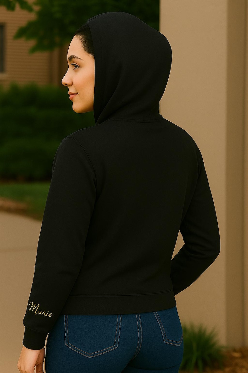 A woman is shown from behind wearing a black hoodie with the hood up. The focus is on the right sleeve, which features the name "Marie" embroidered in elegant gold script near the cuff. She is standing outdoors against a neutral-toned wall and green foliage background, with soft lighting highlighting the clean stitching of the hoodie and the smooth texture of the fabric.