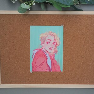 Peut inclure: Un tableau en li&egrave;ge encadr&eacute; avec un dessin d'une personne &eacute;pingl&eacute; dessus. Le dessin repr&eacute;sente une personne aux cheveux blonds et une veste rouge sur un fond turquoise. Le tableau a un cadre en bois clair et est mont&eacute; sur un mur gris.