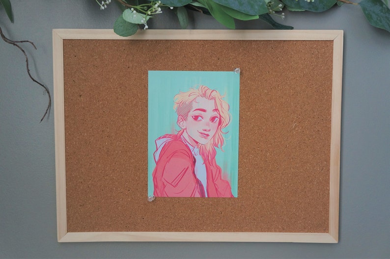 Peut inclure: Un tableau en li&egrave;ge encadr&eacute; avec un dessin d'une personne &eacute;pingl&eacute; dessus. Le dessin repr&eacute;sente une personne aux cheveux blonds et une veste rouge sur un fond turquoise. Le tableau a un cadre en bois clair et est mont&eacute; sur un mur gris.