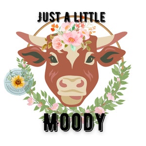 May include: A brown cow with a floral crown, surrounded by a green wreath, with the text "JUST A LITTLE MOODY" in black letters.