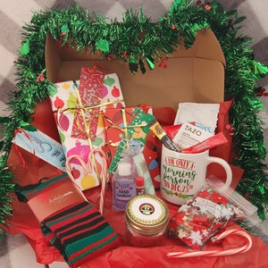 May include: A Christmas-themed gift box filled with wrapped presents, candy canes, socks, a mug, and other festive items. The mug reads "I AM ONLY A morning person ON DEC.25th". The box is decorated with green garland and red tissue paper.