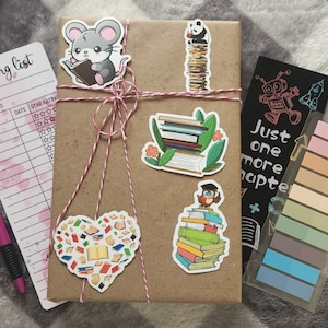 May include: A brown wrapped book with a reading list, a pen, and several book-themed stickers. The stickers include a mouse reading a book, a panda on a stack of books, a pile of books with leaves, an owl on a stack of books, and a heart made of books. The book is tied with twine and has a black bookmark with the text "Just one more chapter".