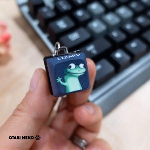 May include: A square, blue keycap with a cartoon lizard design and the word "LIZARD" printed on top. The keycap is held in front of a black keyboard.