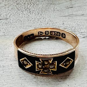 May include: A gold ring with a black enamel band. The ring features a cross design with small, round gemstones and diamond-shaped accents. The ring is marked with the number "9 375".