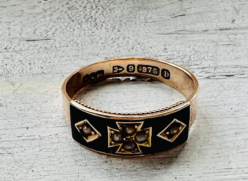 May include: A gold ring with a black enamel band. The ring features a cross design with small, round gemstones and diamond-shaped accents. The ring is marked with the number "9 375".
