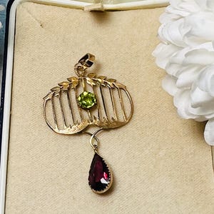 May include: A gold-toned pendant with a cage-like design, featuring a round green gemstone and a teardrop-shaped red gemstone. The pendant is displayed in a vintage jewelry box, showcasing its intricate details and elegant style.