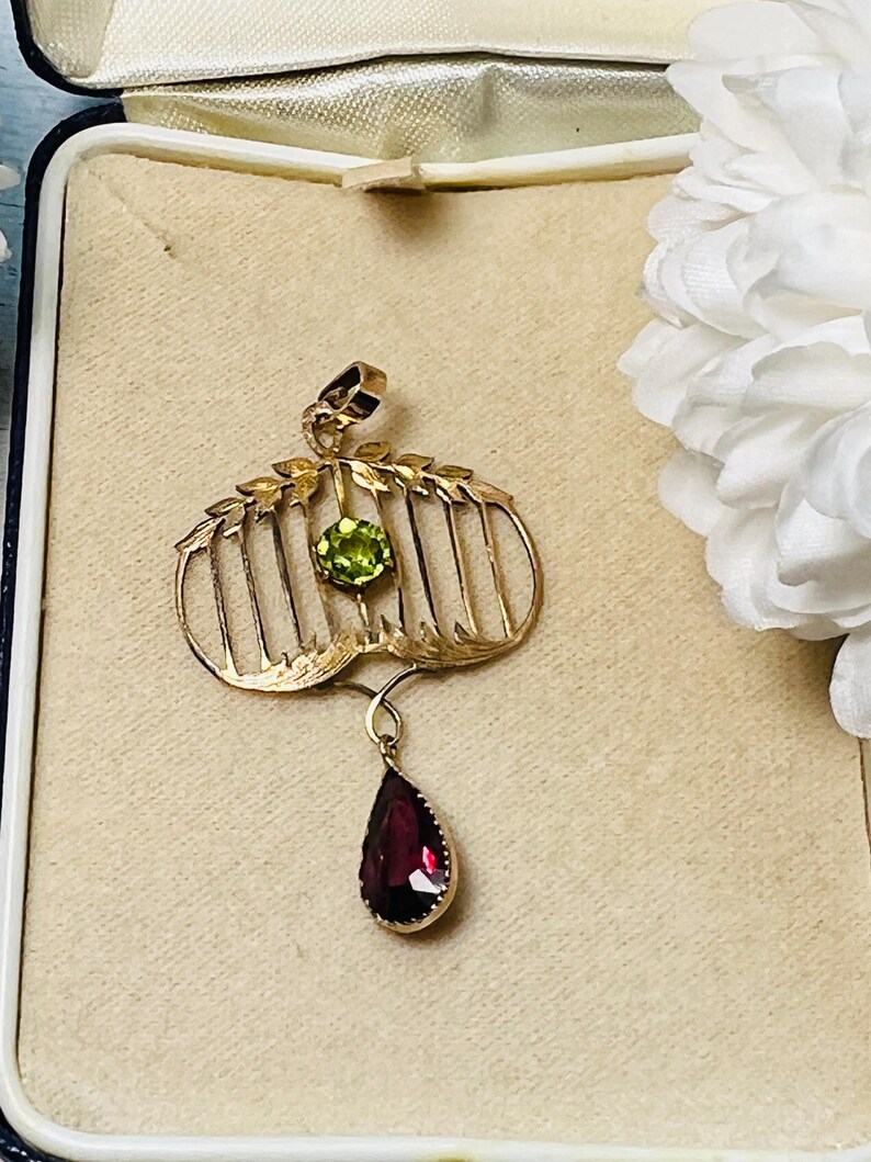 May include: A gold-toned pendant with a cage-like design, featuring a round green gemstone and a teardrop-shaped red gemstone. The pendant is displayed in a vintage jewelry box, showcasing its intricate details and elegant style.