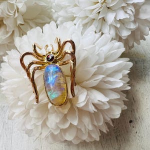 May include: A gold-toned spider brooch with a large, iridescent oval stone at its center. The brooch has a small, red gemstone at the head and is set against a backdrop of white fabric flowers.