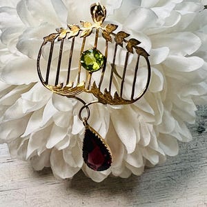May include: A gold-toned vintage brooch with a cage-like design, featuring a round green gemstone and a teardrop-shaped red gemstone pendant. The brooch has leaf accents and vertical bars, set against a white floral background.