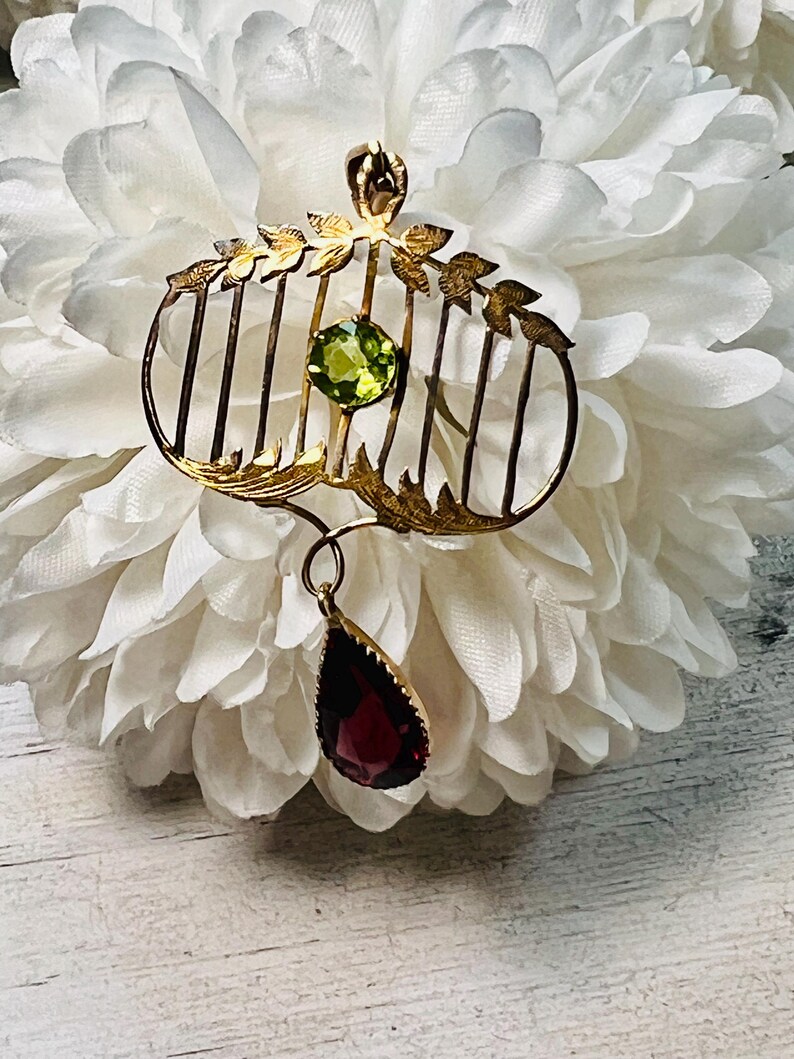 May include: A gold-toned vintage brooch with a cage-like design, featuring a round green gemstone and a teardrop-shaped red gemstone pendant. The brooch has leaf accents and vertical bars, set against a white floral background.