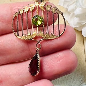 May include: A gold-toned pendant with a cage-like design, featuring a round green gemstone and a teardrop-shaped, dark red gemstone. The pendant has leaf-like accents and vertical bars, creating a vintage aesthetic. The gemstones are the focal point of this jewelry piece.